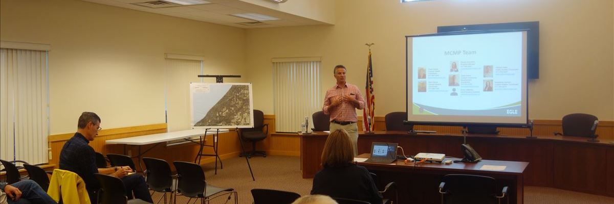 A person addresses people in a room with a screen and many chairs.